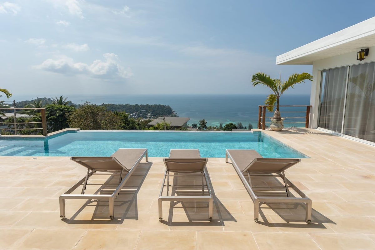 A sunlit patio features three lounge chairs arranged beside a sparkling infinity pool. The scene overlooks Haad Salad Bay, showcasing panoramic sea views and lush greenery under a clear blue sky.