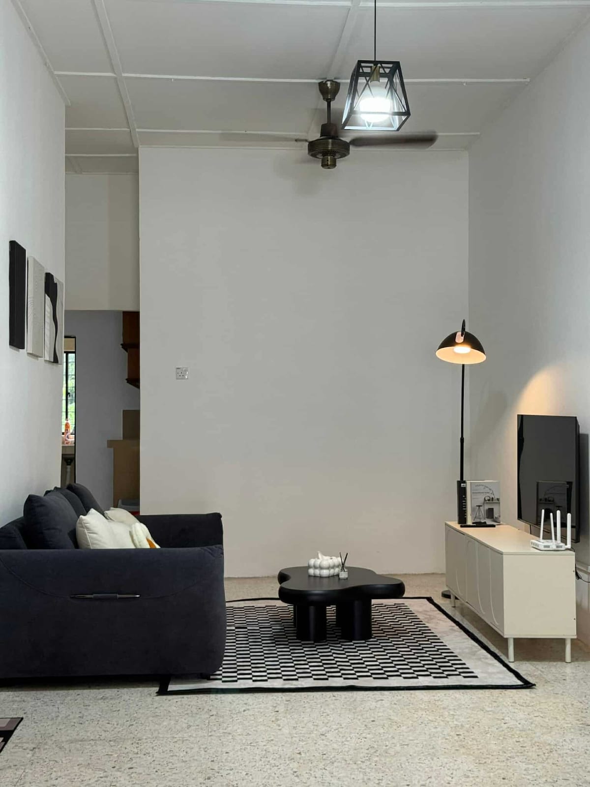 A comfortable living area features a dark sofa with decorative pillows, paired with a low black coffee table. A patterned rug grounds the space, while a floor lamp and wall-mounted television add functionality. High ceilings enhance the airy atmosphere.