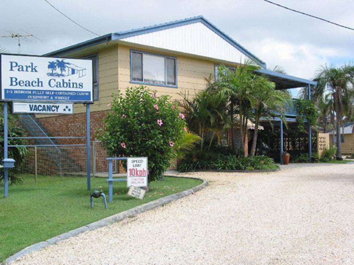 The exterior of the cabin complex is shown, featuring a welcoming sign that indicates 'Park Beach Cabins' and 'Vacancy.' Surrounding greenery and tropical plants enhance the setting, while a gravel path leads to the entrance. The building displays a two-story structure with balconies.