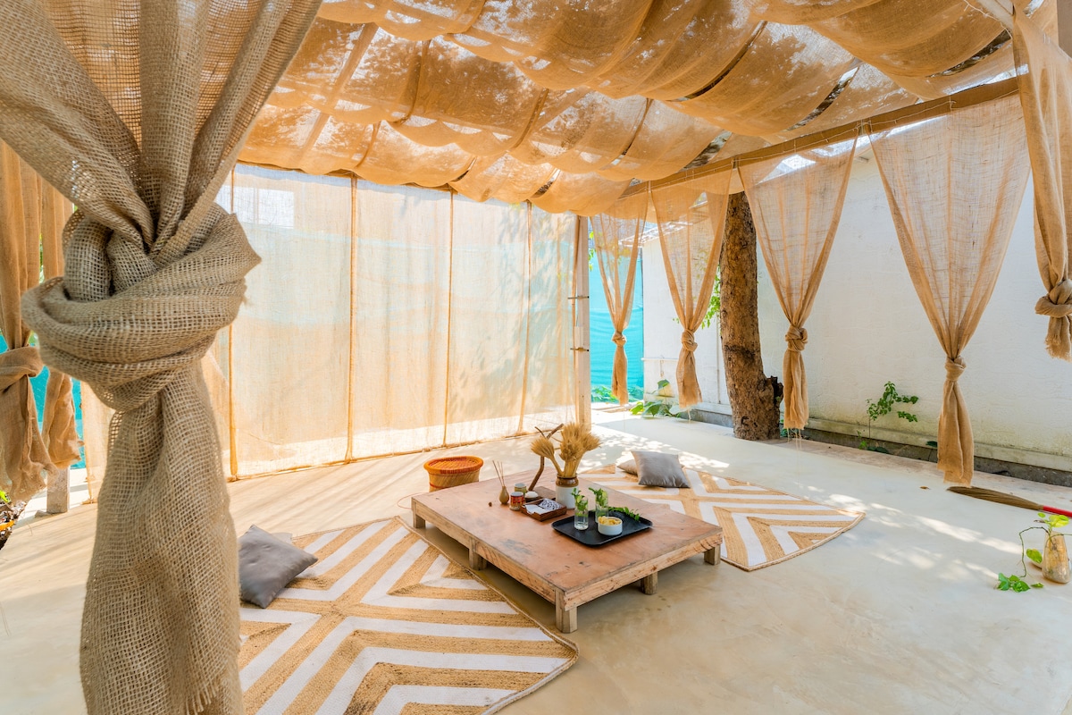 An inviting outdoor relaxation area features a sandy floor covered with a jute mat. Light fabric drapes provide gentle shade, while a low wooden table holds decorative items and drinks. Cushions are placed along the edges, creating a serene atmosphere surrounded by greenery.