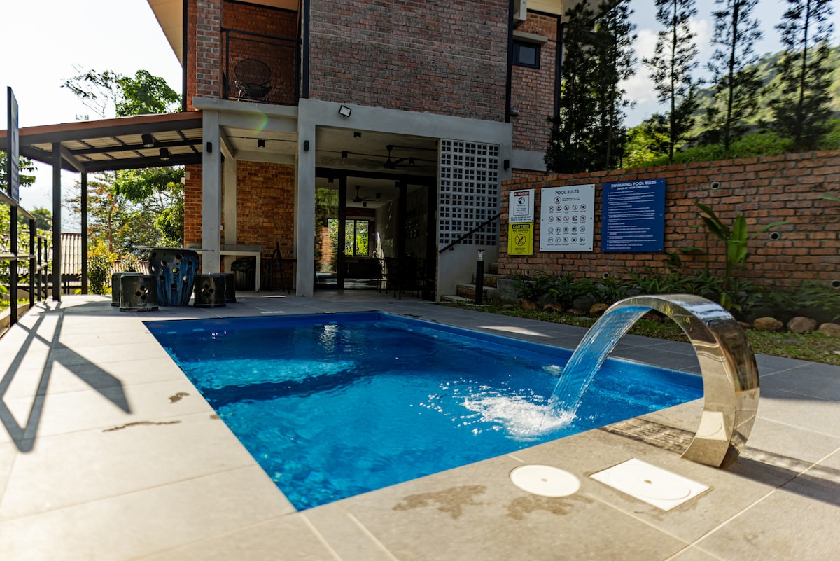 A private pool is featured with a sleek stainless steel water feature, creating a soothing waterfall effect. The pool area is surrounded by tiled flooring and lush greenery, with the villa's brick exterior providing a warm backdrop. Sunlight reflects off the water's surface.