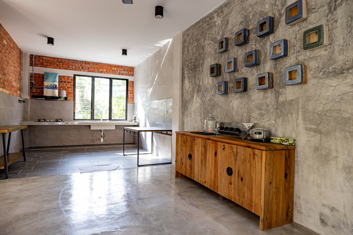 A modern kitchen area features a mix of exposed brick and smooth concrete walls. Natural light streams through large windows, illuminating a sturdy wooden sideboard and dining table. The space is designed with a minimalist aesthetic, promoting a clean and functional environment.