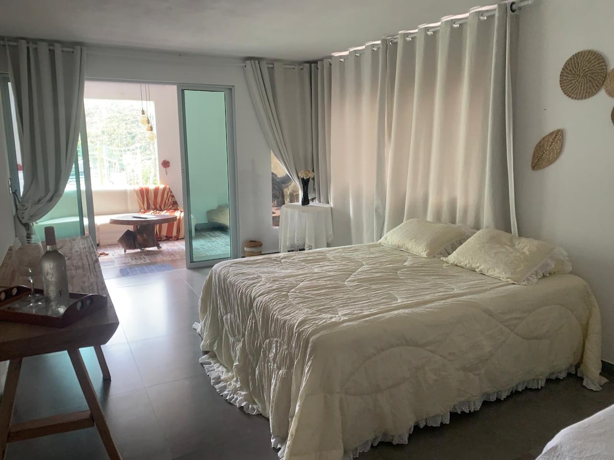 A bright sleeping area is presented with a spacious bed covered in a light quilt. Soft curtains drape across the room, and a sliding door leads to an outdoor space visible in the background. A wooden table and decorative wall elements add subtle detail.