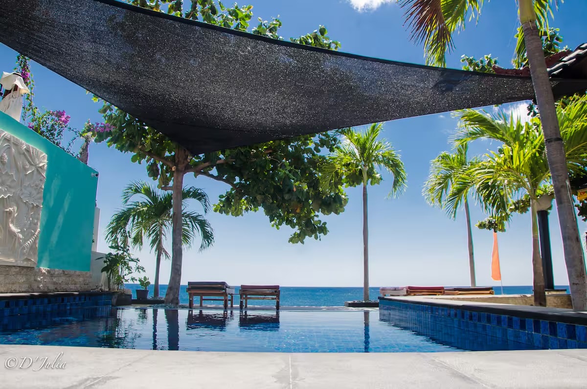 An infinity pool is featured with a backdrop of the ocean, framed by rows of palm trees and vibrant tropical foliage. Sun loungers are positioned nearby for relaxation under a shaded area, creating an inviting space for guests to enjoy the scenic views.