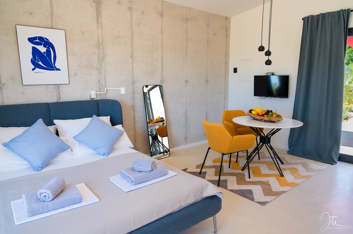 A modern studio apartment features a spacious bed with light blue pillows and neatly arranged towels. A round dining table with yellow chairs sits nearby, complemented by a chevron-patterned rug. A mirror reflects natural light, and a flat-screen TV is mounted on the wall.