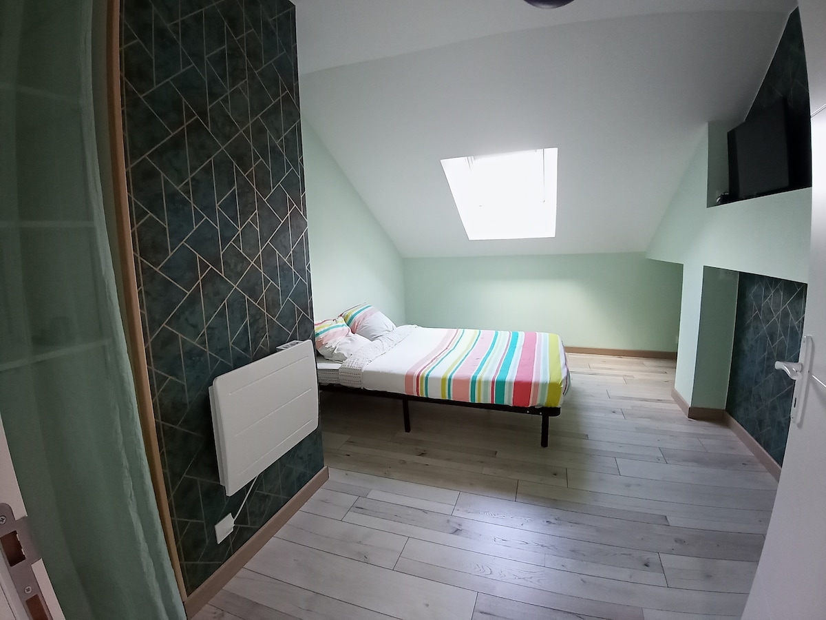 A cozy room is captured showing a spacious bed adorned with colorful striped bedding. Light pours in through a skylight, illuminating the green walls and highlighting the geometric accent wall. A television is mounted on the wall opposite the bed, offering entertainment options.