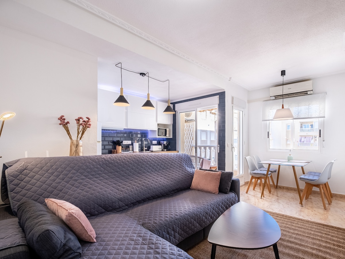 A spacious living area features a comfortable sofa with decorative pillows, accompanied by a coffee table. A dining table for four is located nearby, with natural light streaming from the large windows. The open kitchen area is visible in the background, showcasing modern appliances.