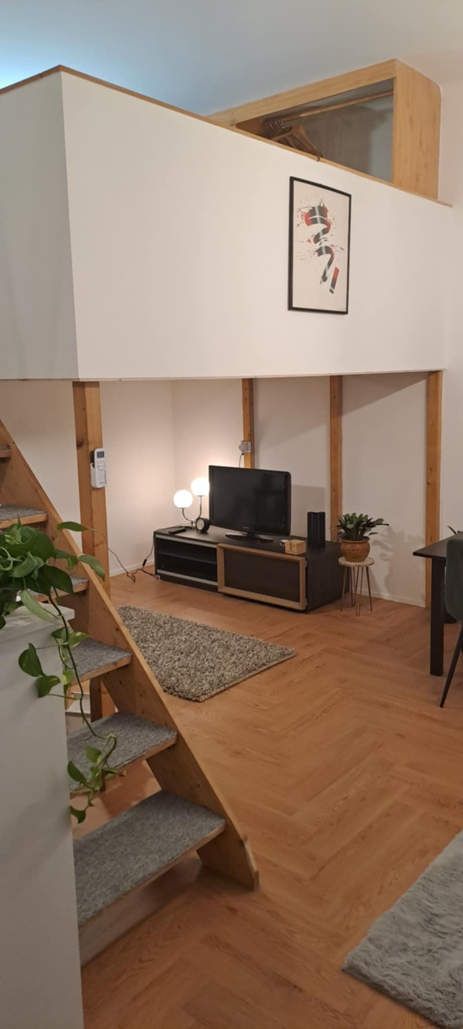 The living area features wooden beams and a warm wooden floor. A television is placed on a low black cabinet, accompanied by decorative plants. A small table and chair are visible nearby, with gentle lighting provided by a lamp. Stairs lead to the lofted sleeping area.