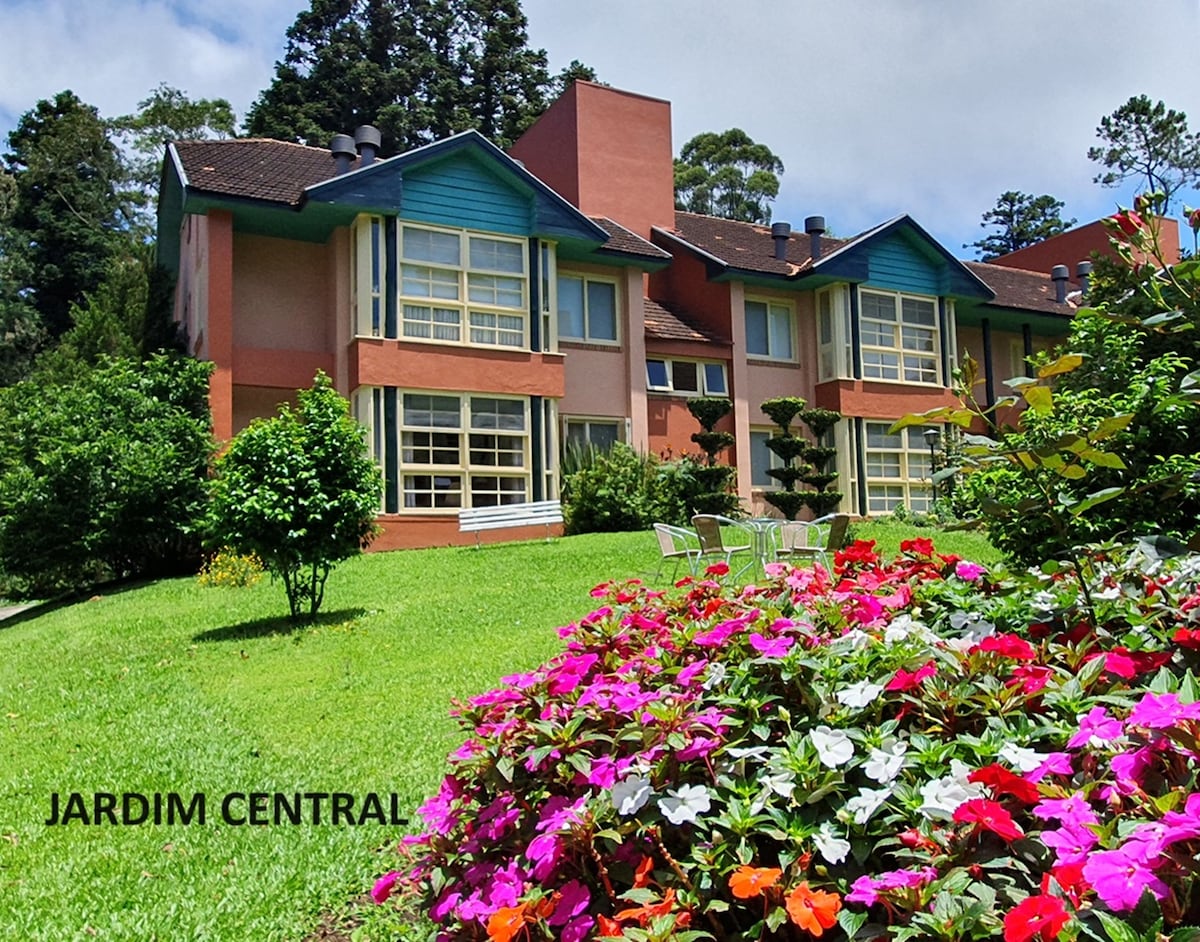 A charming building is set within a lush green landscape, featuring vibrant flower beds in the foreground. The structure showcases large windows that offer a view of the surroundings, with well-manicured grass and shrubs adding to the appeal of the area.