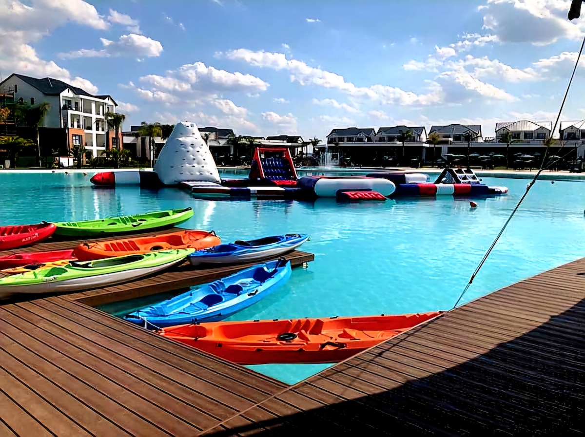 A vibrant lagoon swimming pool is surrounded by colorful kayaks in greens, blues, and oranges, lined along a wooden dock. Inflatable water play structures are visible on the glistening water, under a partly cloudy sky that adds brightness to the scene.