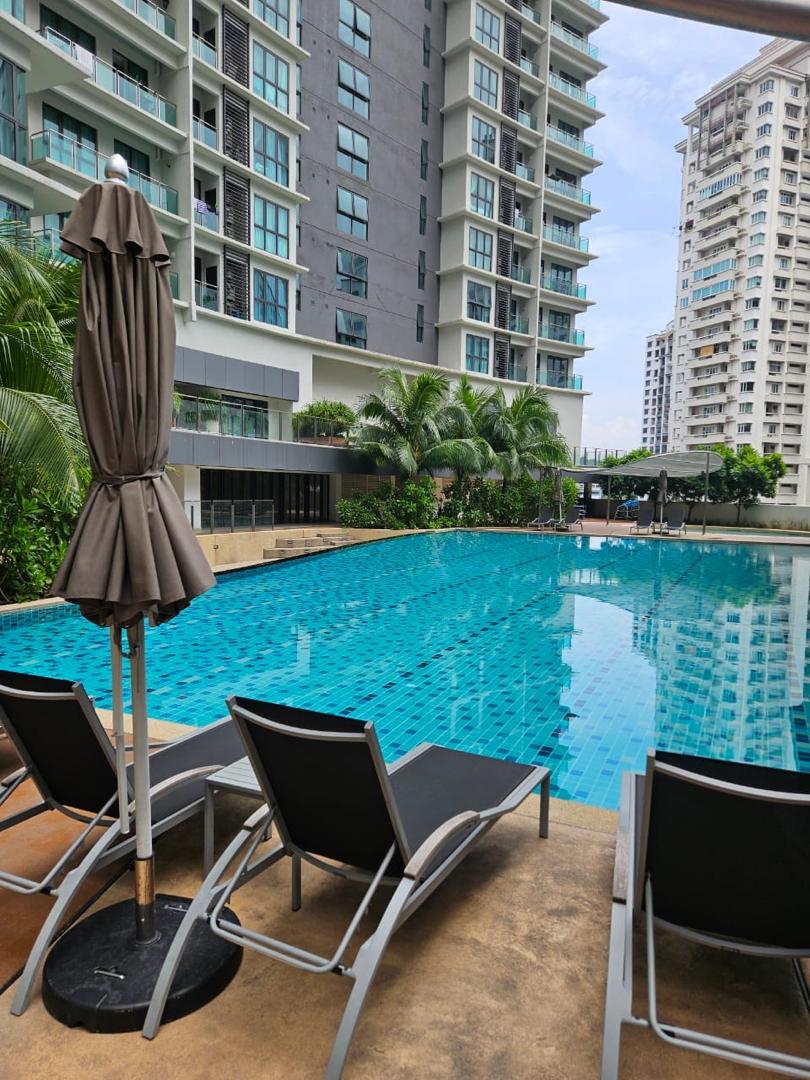 A serene swimming pool is surrounded by lush greenery and modern apartment buildings. Lounge chairs are positioned nearby, with a sun umbrella adding shade. The clear blue water reflects the surrounding structures and sky, creating a calming environment for relaxation.