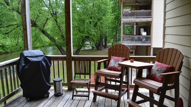 A wooden deck is viewed, featuring two rocking chairs with decorative pillows and a small table. A grill is positioned nearby, while lush greenery and the river can be seen in the background, creating a relaxed outdoor setting.