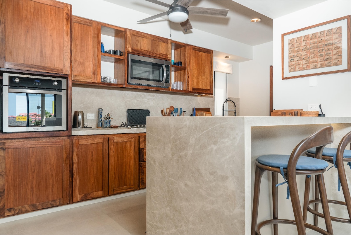 A modern kitchen is equipped with wooden cabinetry and granite countertops. Appliances include a built-in oven, microwave, and kettle. Bar seating is available at the island, featuring three wooden stools. A ceiling fan provides ventilation, and large windows offer natural light.