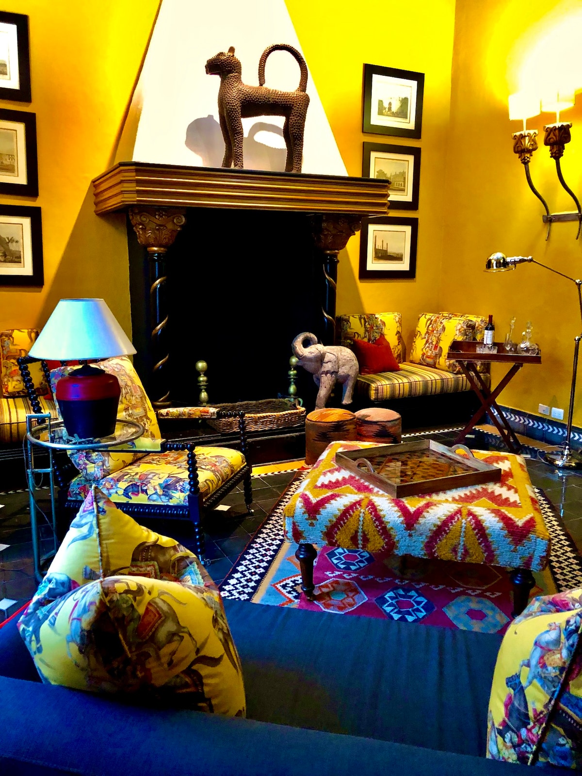 A vibrant sitting room features saffron yellow walls, a decorative fireplace, and various ornate pillows on the seating. A patterned coffee table sits at the center, surrounded by stylish armchairs. Artistic accents, including framed wall art and a ceramic elephant, add character to the space.