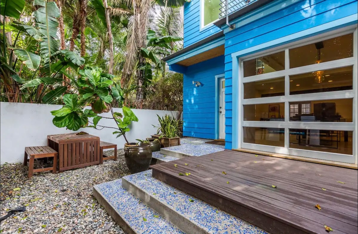 A vibrant blue exterior complements a welcoming entrance, framed by lush greenery. The outdoor area features stone steps leading to a wooden deck, along with a seating area made of natural wood. Decorative pots with greenery add to the inviting ambiance.