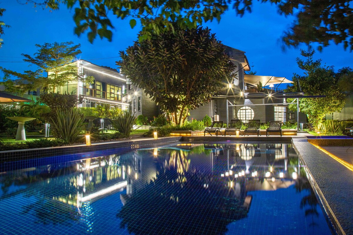 An outdoor pool is surrounded by lush greenery and illuminated by soft lighting. Lounge chairs are placed along the pool’s edge, and a spacious building is visible in the background. The scene conveys a peaceful evening ambiance, enhanced by the reflections on the water.