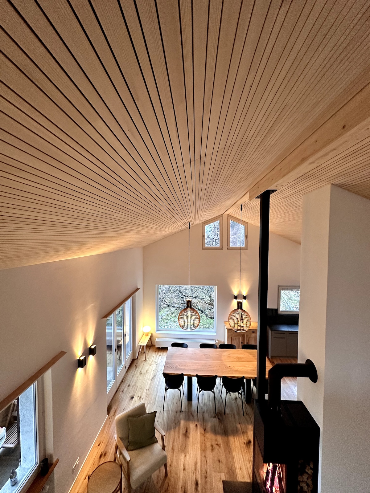A spacious living area features a high wood-paneled ceiling. Natural light enters through large windows, illuminating a modern dining table surrounded by chairs. Two pendant lights hang over the table, while a stove adds warmth to the ambiance. The open layout connects to the kitchen area.