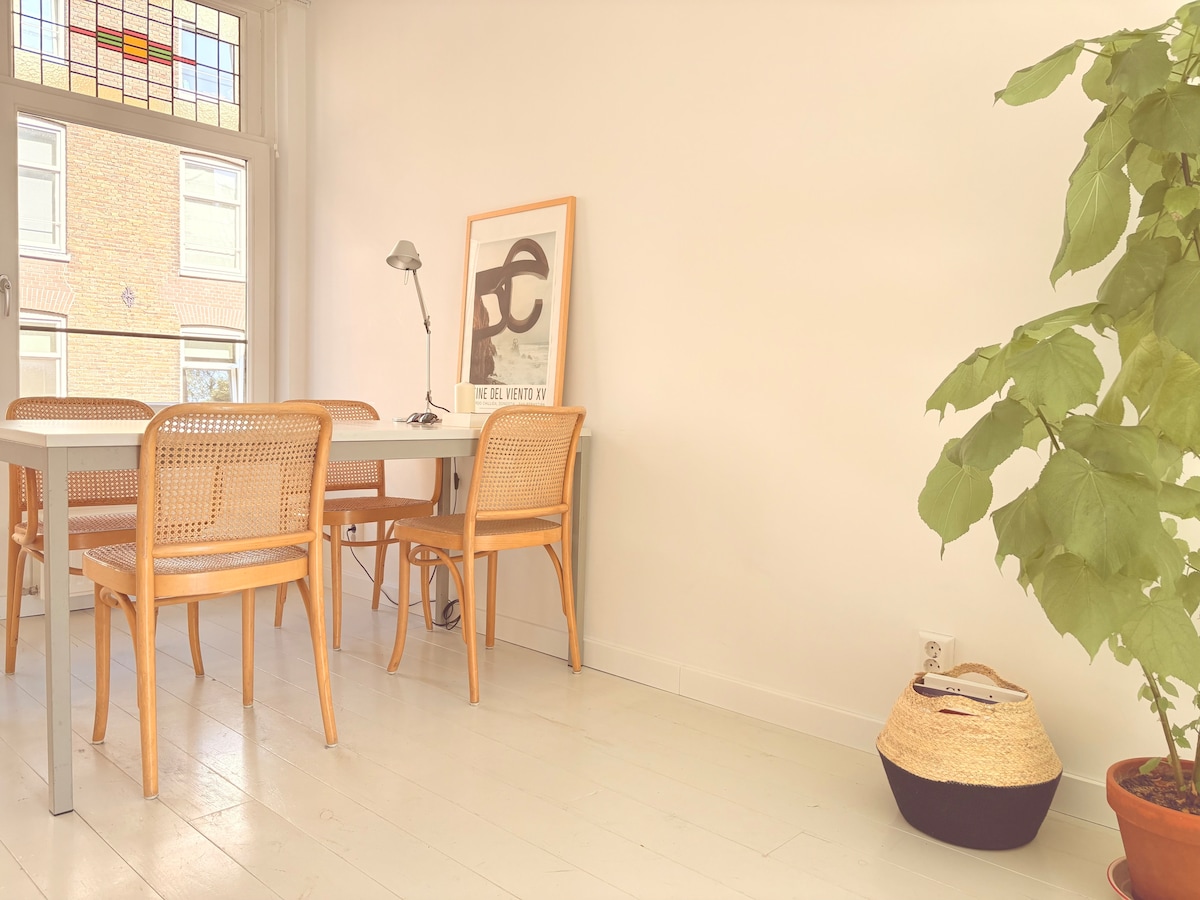 A dining area features a light-colored table surrounded by four wooden chairs with woven backs. A modern lamp stands on the table. A large window allows natural light to fill the space, while a decorative plant in a terracotta pot adds a touch of greenery.