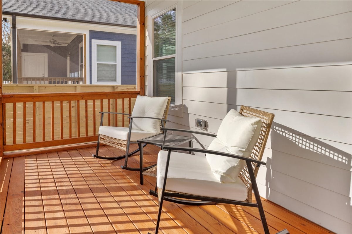 A screened-in porch features two comfortable seating chairs positioned around a glass-top table. Natural light filters through the slats, casting shadows on the wooden flooring. The adjoining area offers views of the outside scenery through the screens.
