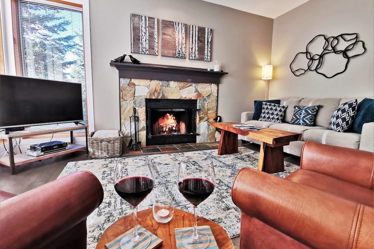 A cozy living area features a stone fireplace with a welcoming fire, surrounded by comfortable seating. Two leather chairs face a rustic coffee table adorned with glasses of red wine. A flat-screen TV sits on a media console, with natural light filtering through the windows.