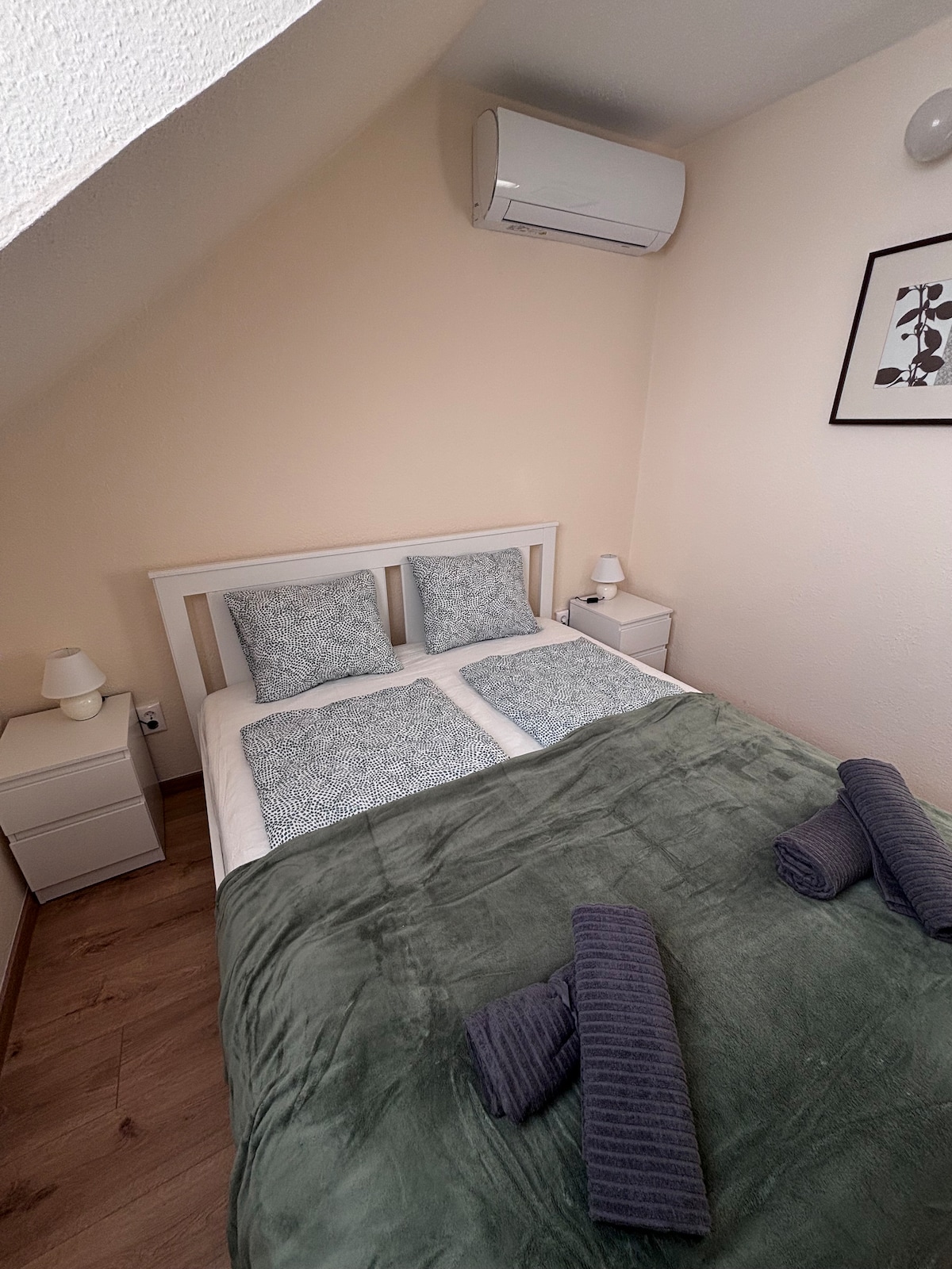 A cozy sleeping area features a spacious bed with light bedding and two patterned pillows. A soft green blanket is draped across the bed, accompanied by two rolled towels. Two bedside tables with lamps are positioned on either side, and an air conditioning unit is mounted on the wall.