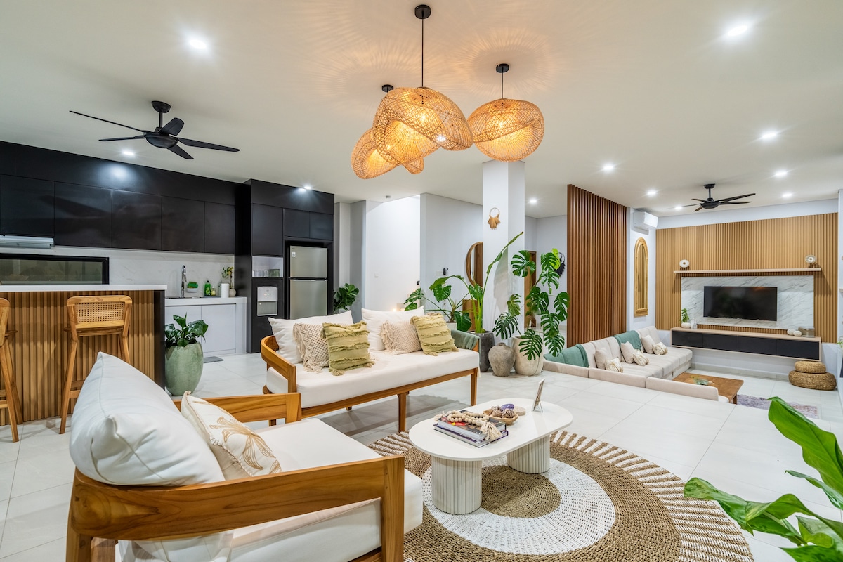 The living area features a modern open-plan design with a comfortable seating arrangement, accentuated by natural materials like wood and rattan. Soft lighting emanates from stylish pendant lamps. Lush greenery enhances the serene ambiance, while a neutral color palette creates a sense of calm.