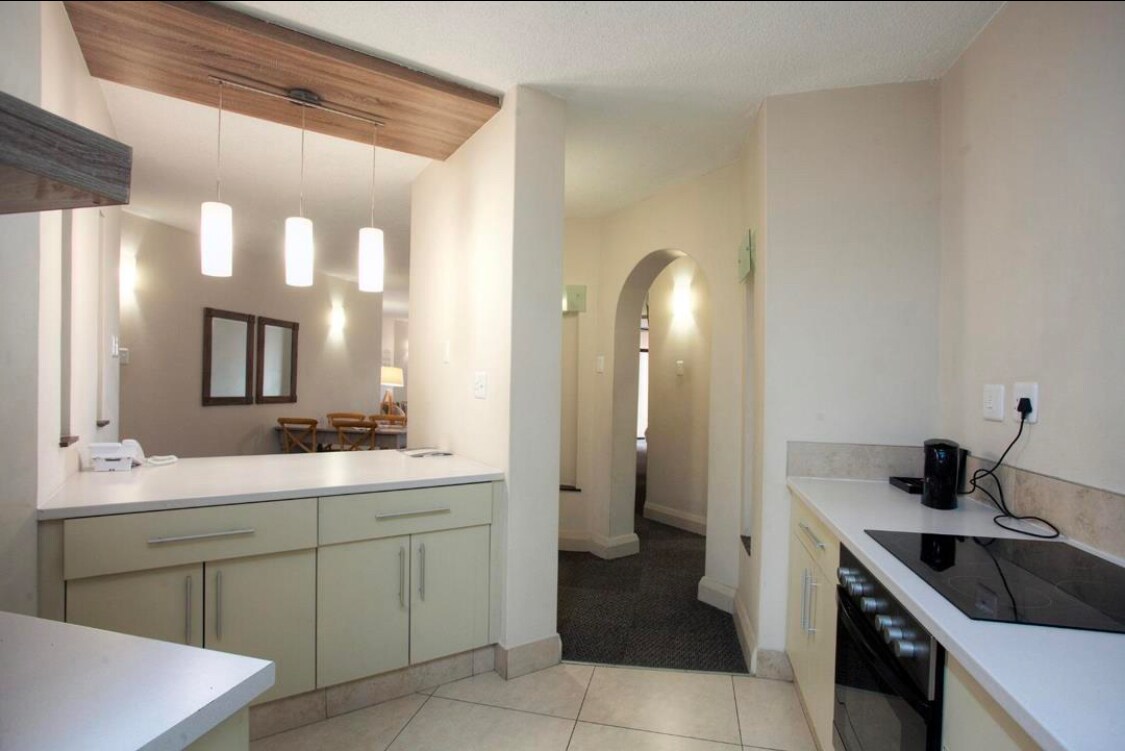 A modern kitchen is displayed with light-colored cabinets and a spacious countertop. Pendant lighting illuminates the area. An arched entryway leads to a softly lit hallway, and a dining area is visible in the background. Appliances, including a coffee maker, are neatly arranged.