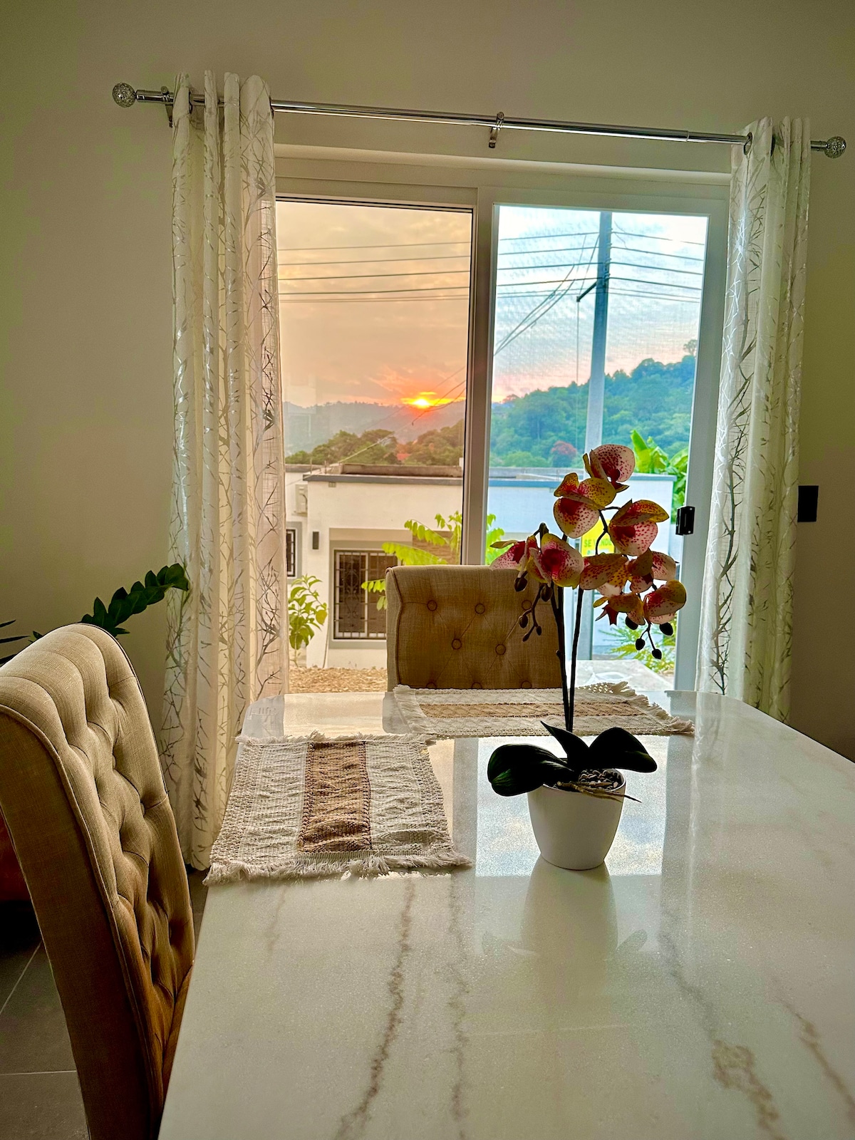 A dining area features a marble table with soft, cushioned chairs. A potted orchid sits in the center. Sheer curtains frame a window showcasing a sunset view over the mountains, illuminating the space with warm hues.