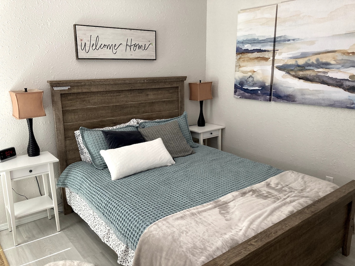 A welcoming bedroom features a wooden bed frame with a light blue and white comforter. Two nightstands with lamps flank the bed. Above, wall art in soft hues of blue and gray adds a serene touch. A sign reading 'Welcome Home' enhances the atmosphere.
