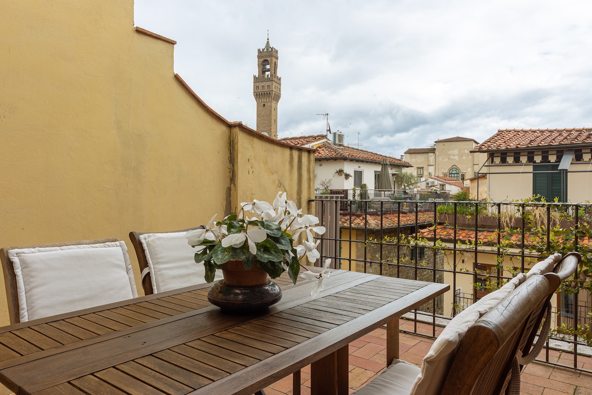 A spacious terrace is presented with a wooden dining table surrounded by comfortable seating. A planter filled with white flowers is centered on the table, with a backdrop of historic buildings and a clock tower in the distance under a partly cloudy sky.