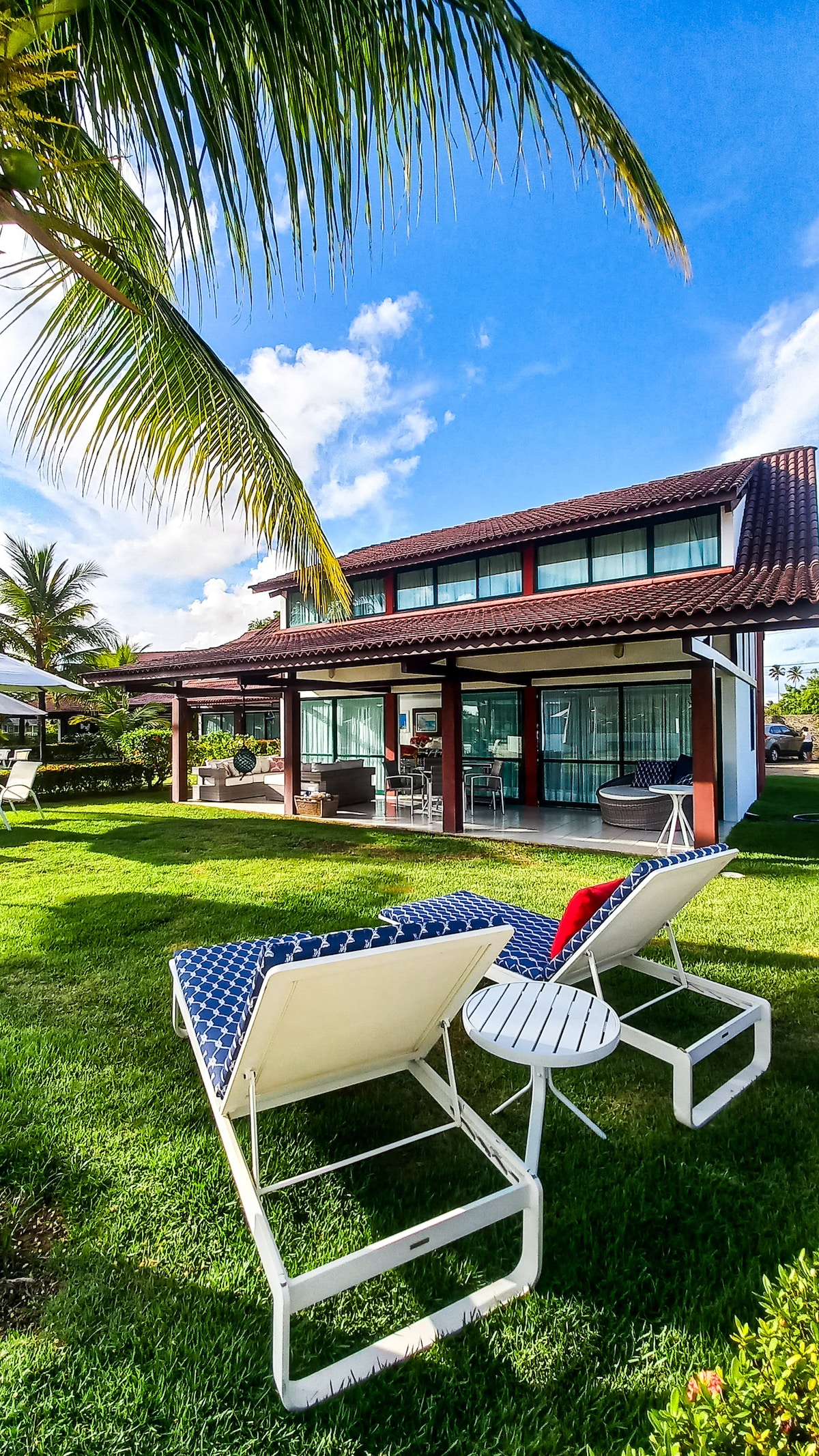 A spacious outdoor area is depicted, featuring lounge chairs positioned on a lush green lawn. A two-story house is visible in the background, framed by palm trees. Large windows allow natural light to illuminate the inviting space, which includes a covered patio area.