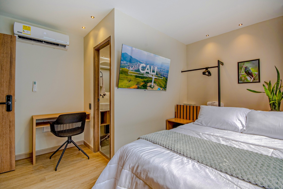 A modern studio features a spacious bed with soft linens and a view of Cali displayed on the wall-mounted Smart TV. A small desk with a chair is positioned to the left, while a plant adds greenery to the space. The bathroom entrance is visible in the background.