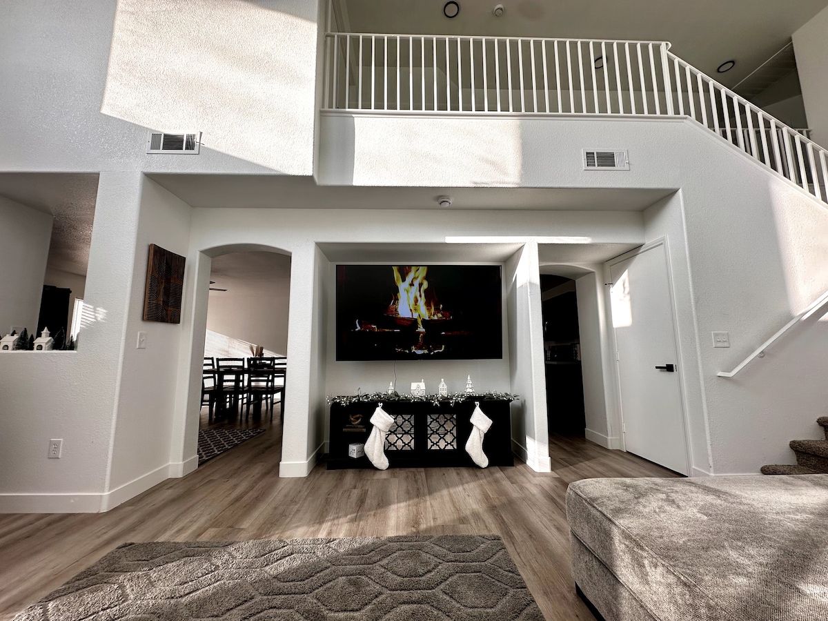The living area is designed with a large television mounted on a wall, featuring a cozy fireplace display. A sectional sofa is placed on a textured area rug, while holiday stockings are hung on a decorative console beneath the TV. Natural light flows in from nearby windows.