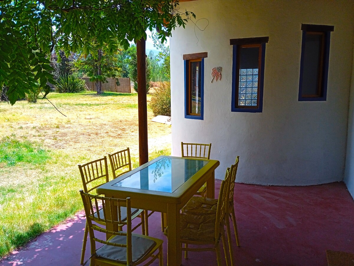 An outdoor dining area is shown with a large yellow table surrounded by eight chairs. The setting is shaded by leafy trees, with a grassy landscape visible in the background. Large windows allow natural light to enter the adjacent space.