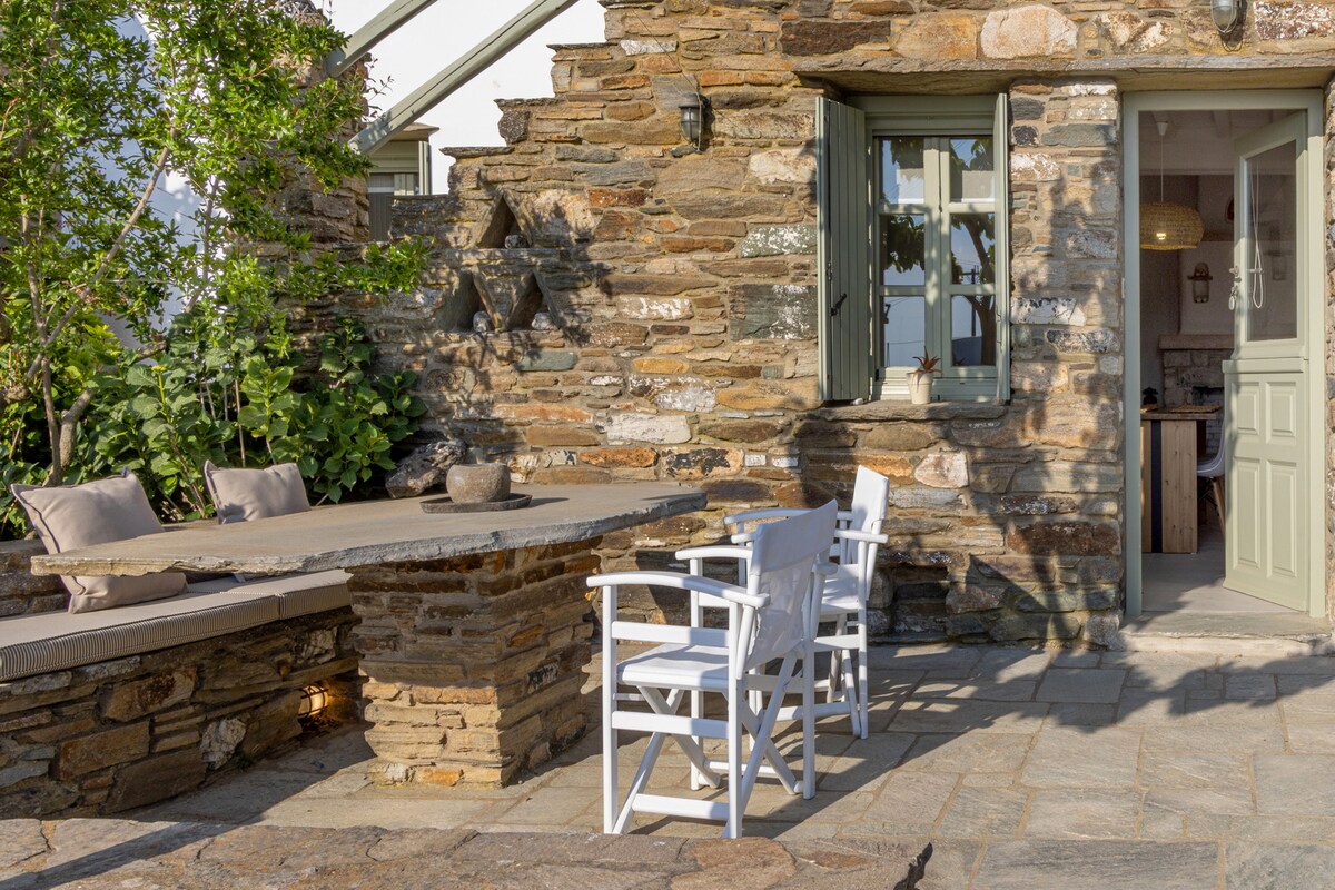 An outdoor seating area features two white chairs positioned around a stone table, surrounded by lush greenery. Textured stone walls and large, open doors lead to the indoor space, allowing natural light to flow through. A peaceful and inviting setting is created.