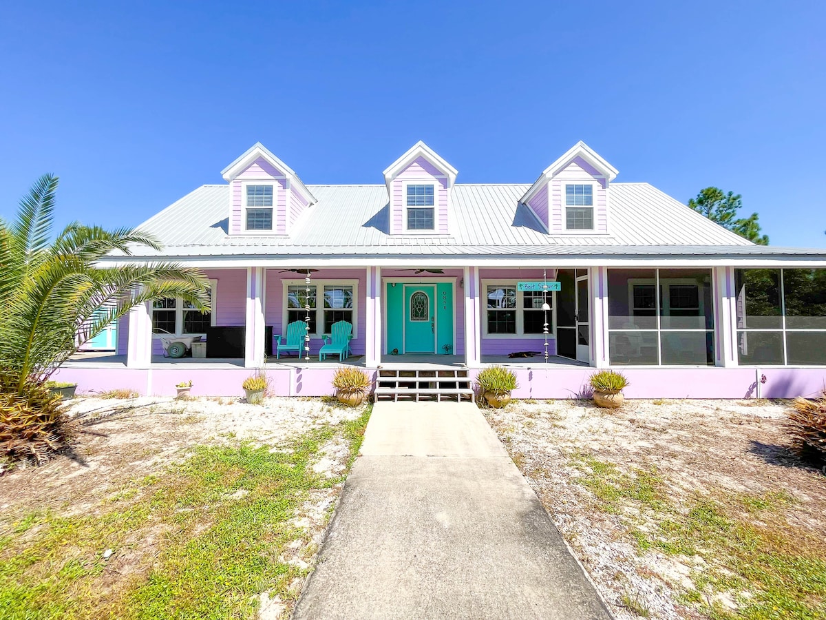 A charming coastal home is presented with a pastel purple exterior and a light blue front door. The covered porch features a mix of seating options, while landscaped areas include low shrubs and palm trees. A clear blue sky is visible overhead.