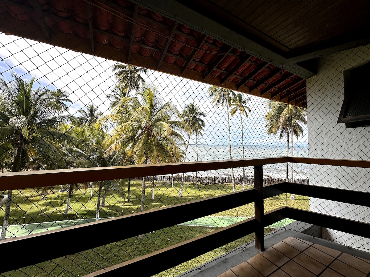 A balcony view is captured, highlighting lush green grass and palm trees swaying gently along the shoreline. The ocean is visible in the distance beneath a clear sky, while a safety netting frames the open space, ensuring a secure experience.