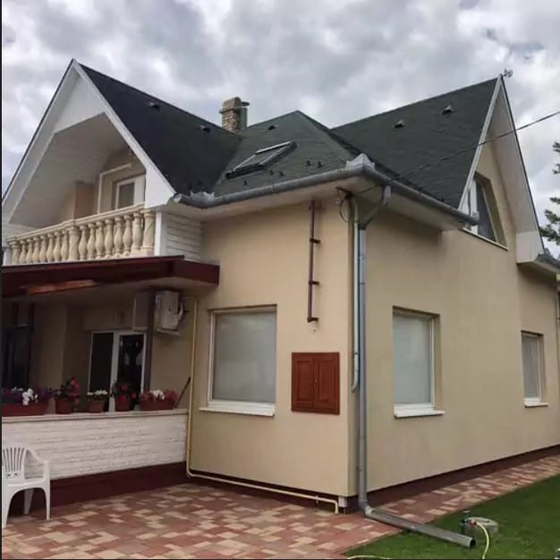 The guesthouse exterior is characterized by a combination of light colors and a sloped roof. Decorative balcony railings are visible above the entrance, which features a small porch with flower pots. Large windows provide ample natural light to the interior.