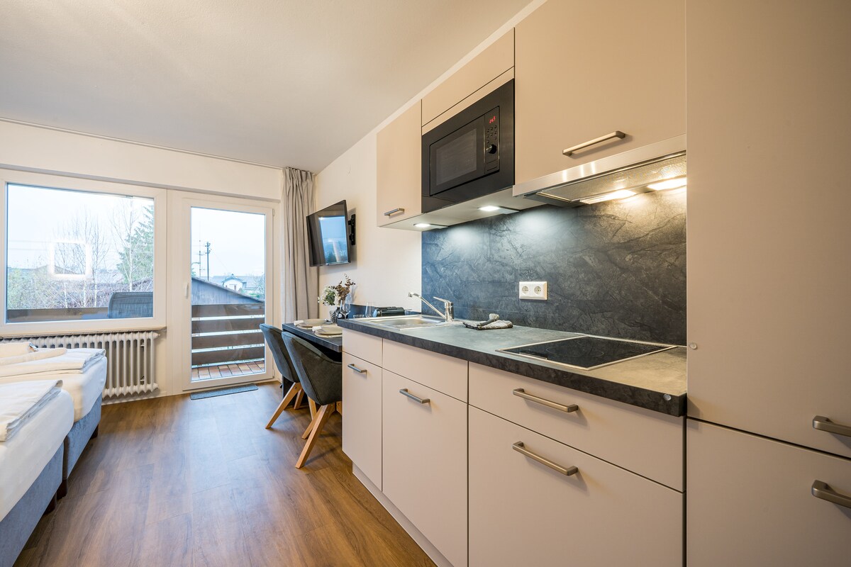 A modern kitchen area features sleek cabinetry and a microwave above the countertop. Natural light enters through large windows, illuminating the space. A seating area with dark chairs and a television provides a comfortable environment, with a glimpse of outdoor scenery visible through the glass doors.