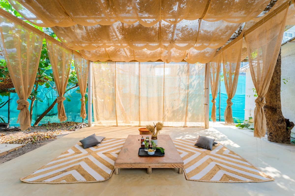 A shaded outdoor seating area is presented, featuring a wooden low table at its center surrounded by patterned floor mats. Soft pillows are arranged around the table, while sheer drapes provide privacy and filter natural light, contributing to a serene atmosphere.