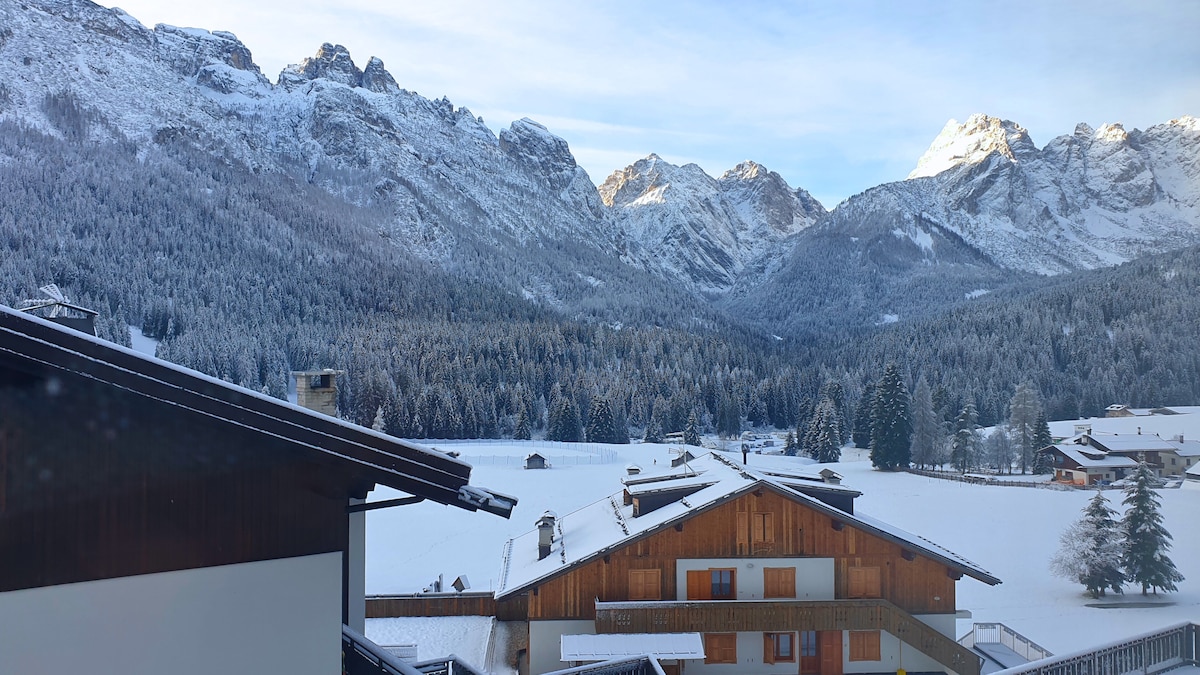 A scenic view showcases the snow-covered mountains and surrounding forest, blanketed in white. Roofs of nearby chalets can be seen below, with the expansive snowy landscape stretching towards the mountains under a clear sky.