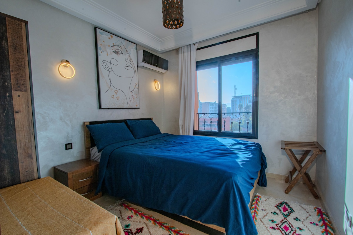 A modern bedroom features a spacious bed with blue bedding, complemented by artistic wall decor. Natural light enters through the large window with a city view, while a bedside table and a small stool add functionality to the space. A cozy area rug is present on the floor.