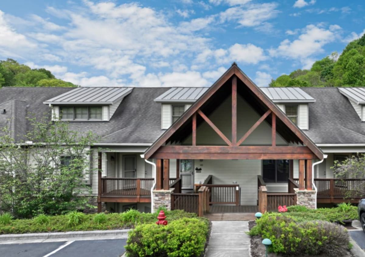 The exterior view displays a charming building with a prominent peaked roof and wooden features. Lush greenery surrounds the entrance, complemented by a stone walkway leading to the entry. The sky above is bright and partly cloudy, enhancing the peaceful ambiance of the neighborhood.