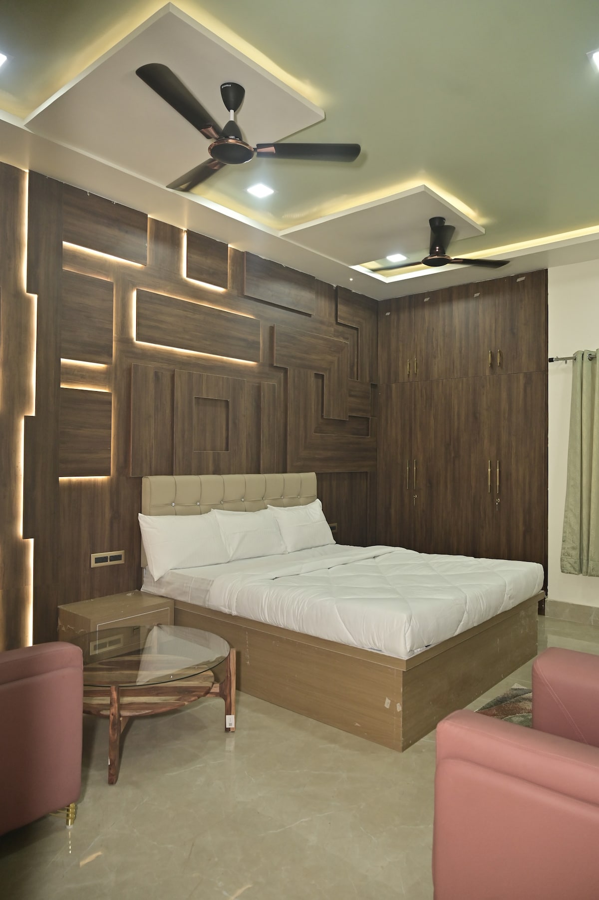 A comfortable room is equipped with a spacious bed featuring a light-colored headboard. The wood-paneled wall design adds texture, complemented by soft lighting. Two armchairs in a muted pink hue are positioned near a small glass table, creating an inviting seating area.