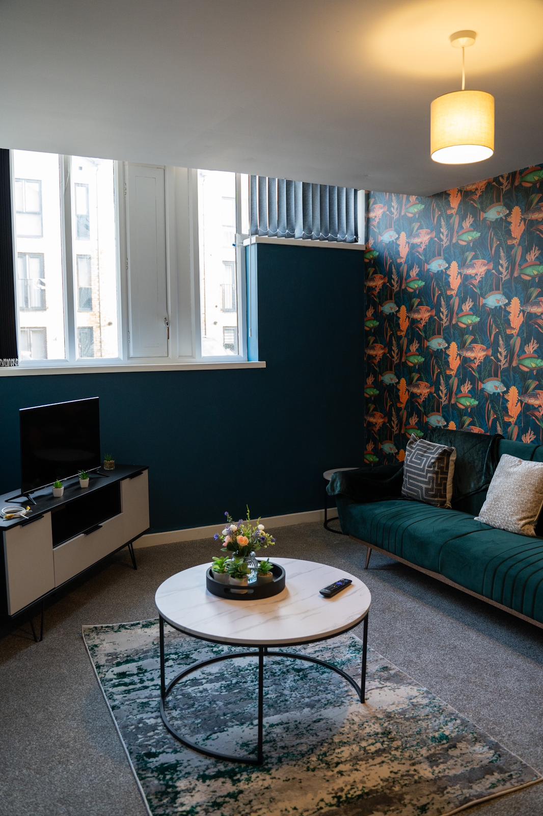 A living area is furnished with a green sofa and a patterned accent wall featuring vibrant foliage. A round coffee table, adorned with a small floral arrangement, is centrally placed. Natural light enters through large windows, complemented by a soft overhead light.