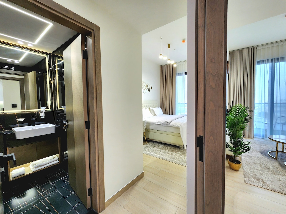 An entrance to the apartment is displayed, showing a view of the bathroom and the bedroom beyond. The bathroom features modern finishes and a stylish sink. In the bedroom, a spacious bed is visible, along with large windows covered by light curtains, allowing natural light to fill the space.