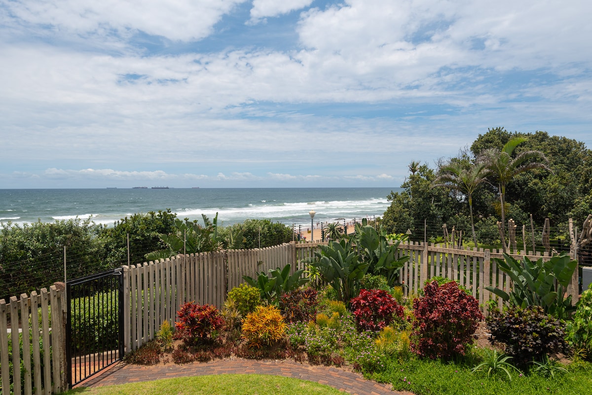 A view of the ocean is presented from the terrace, featuring gentle waves and a sandy beach in the distance. Lush greenery and colorful flowers line the garden, framed by a wooden fence, creating a serene outdoor space inviting relaxation.
