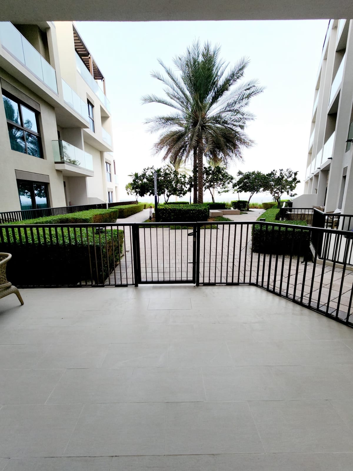 The image showcases a serene outdoor area featuring manicured greenery and palm trees. A gated entrance is visible, leading into a spacious courtyard. The surrounding architecture includes modern balconies and large windows, reflecting a tranquil setting.