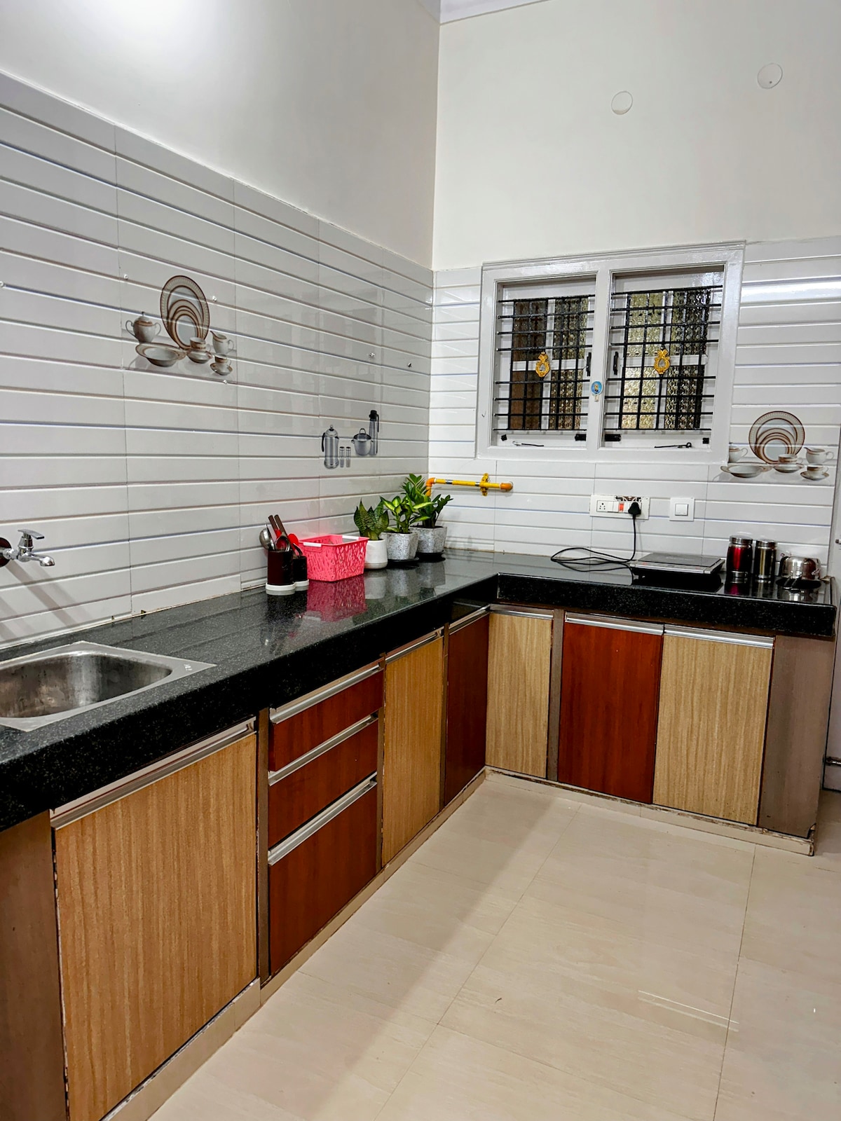 A well-organized kitchen features sleek black granite countertops and wooden cabinetry in contrasting shades. Essential cooking utensils, plants for added freshness, and a stainless steel sink are visible, along with a window allowing natural light in while providing ventilation.