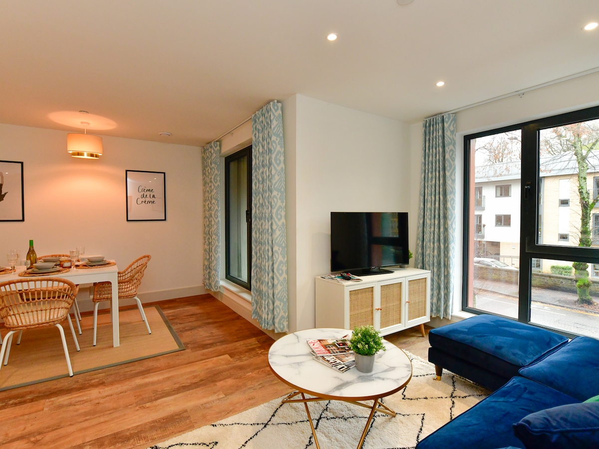 A modern open-plan living area features a comfortable blue sofa and a marble coffee table. Natural light fills the space through large windows, accompanied by stylish curtains. A dining table set with chairs is visible in the background, creating a welcoming atmosphere for gatherings.